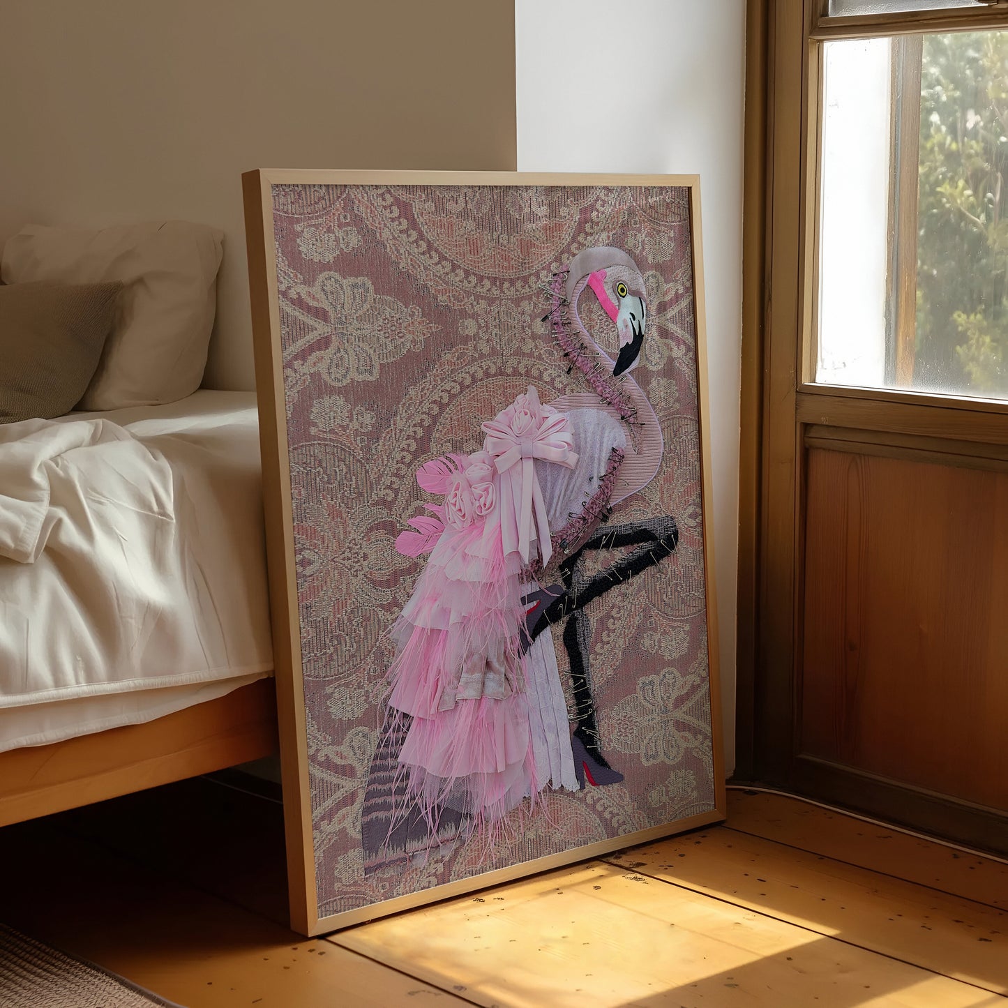 A framed art print featuring a pink flamingo on a patterned background, leaning against a bed in a sunlit bedroom.