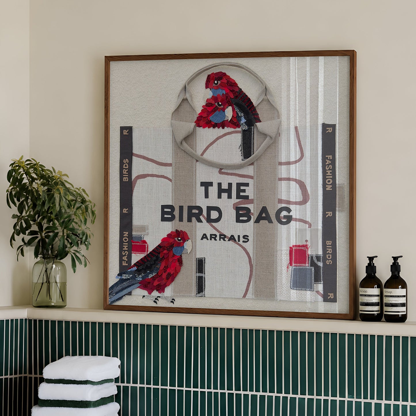 A framed art print featuring two red and blue parrots with the text 'THE BIRD BAG ARRAIS' is displayed on a bathroom shelf above a green and white striped tile wall. The print is styled with a wooden frame, flanked by a potted plant on the left and two dark bottles on the right.