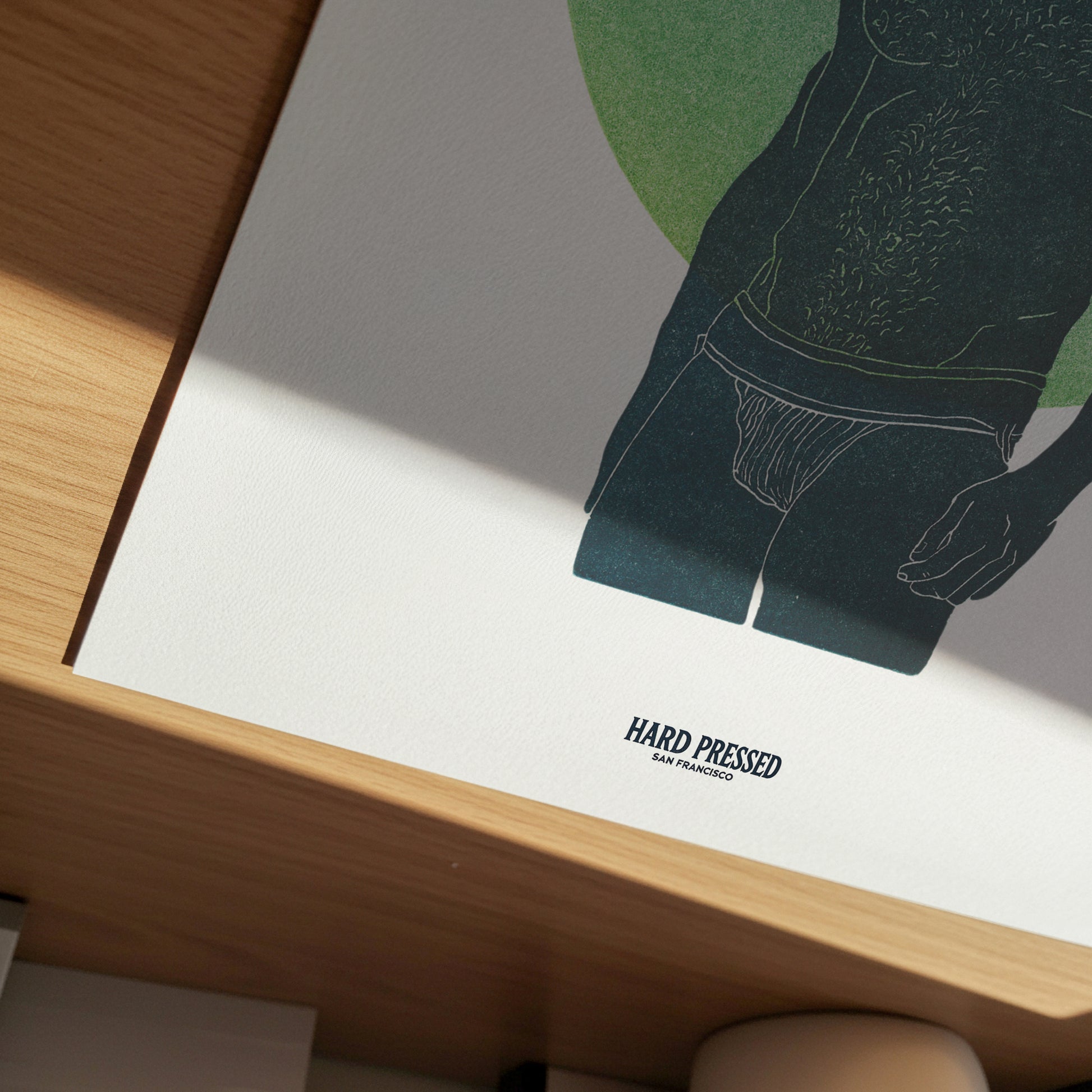 A close-up of a printed poster or art print resting on a wooden surface. The print features a minimalist illustration of a male torso wearing underwear, with a large green circle in the background and the text 'HARD PRESSED SAN FRANCISCO' near the bottom.