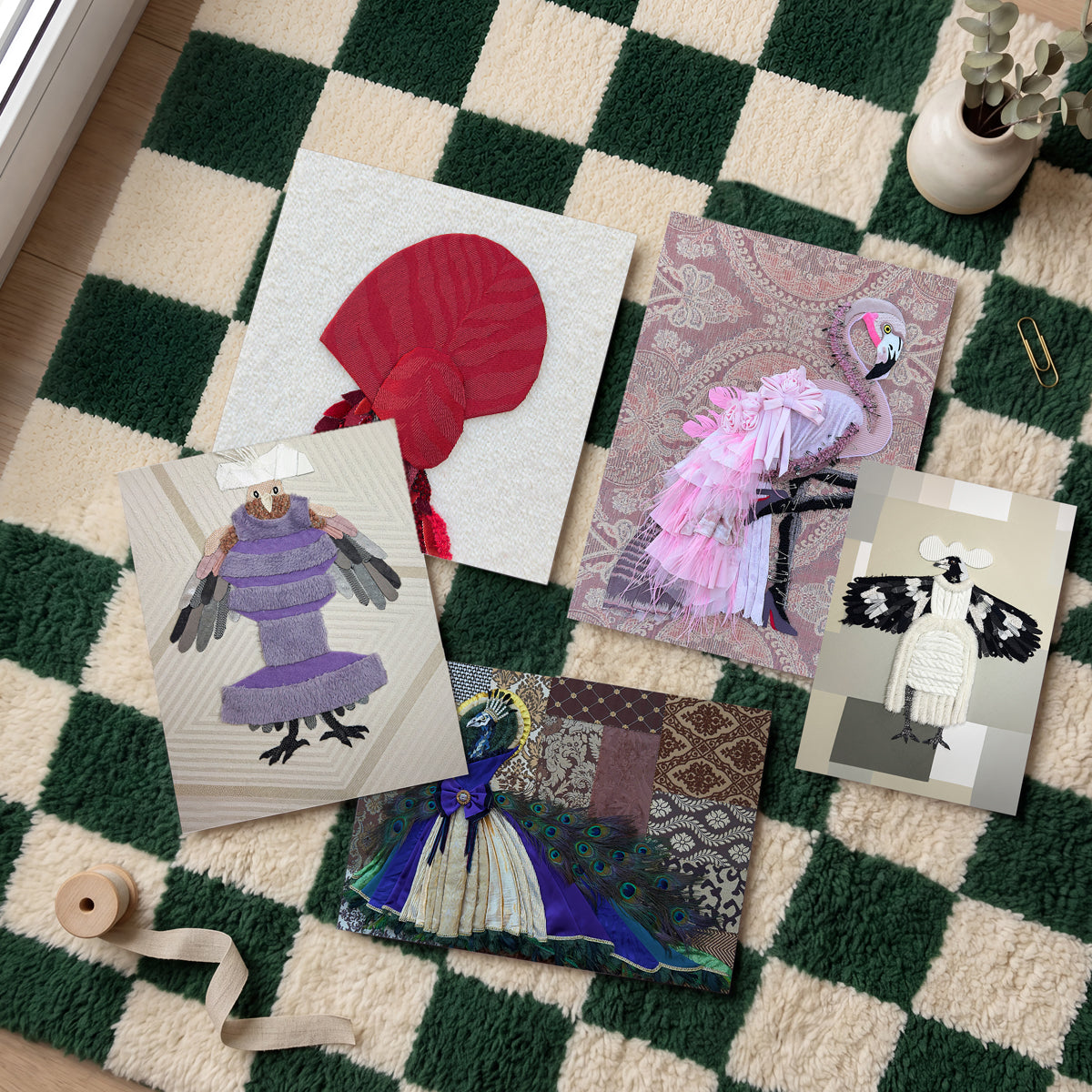 Five handmade art cards featuring whimsical bird and animal illustrations are arranged on a green and cream checkered rug. The cards showcase various colorful patchwork designs, including a red bird, a pink flamingo, a purple bird, a blue bird, and a black-and-white bird, styled with a spool of ribbon and a small potted eucalyptus plant.