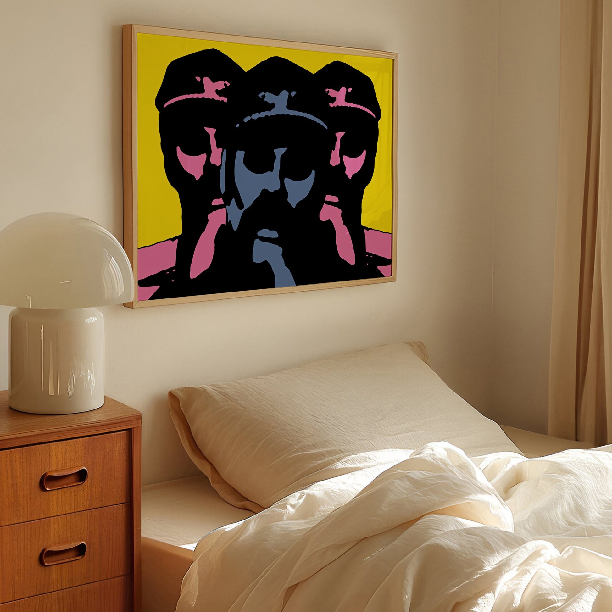 A framed pop art print featuring three stylized faces in black, blue, and pink on a bright yellow background, hanging on a beige wall above a bed in a softly lit bedroom.