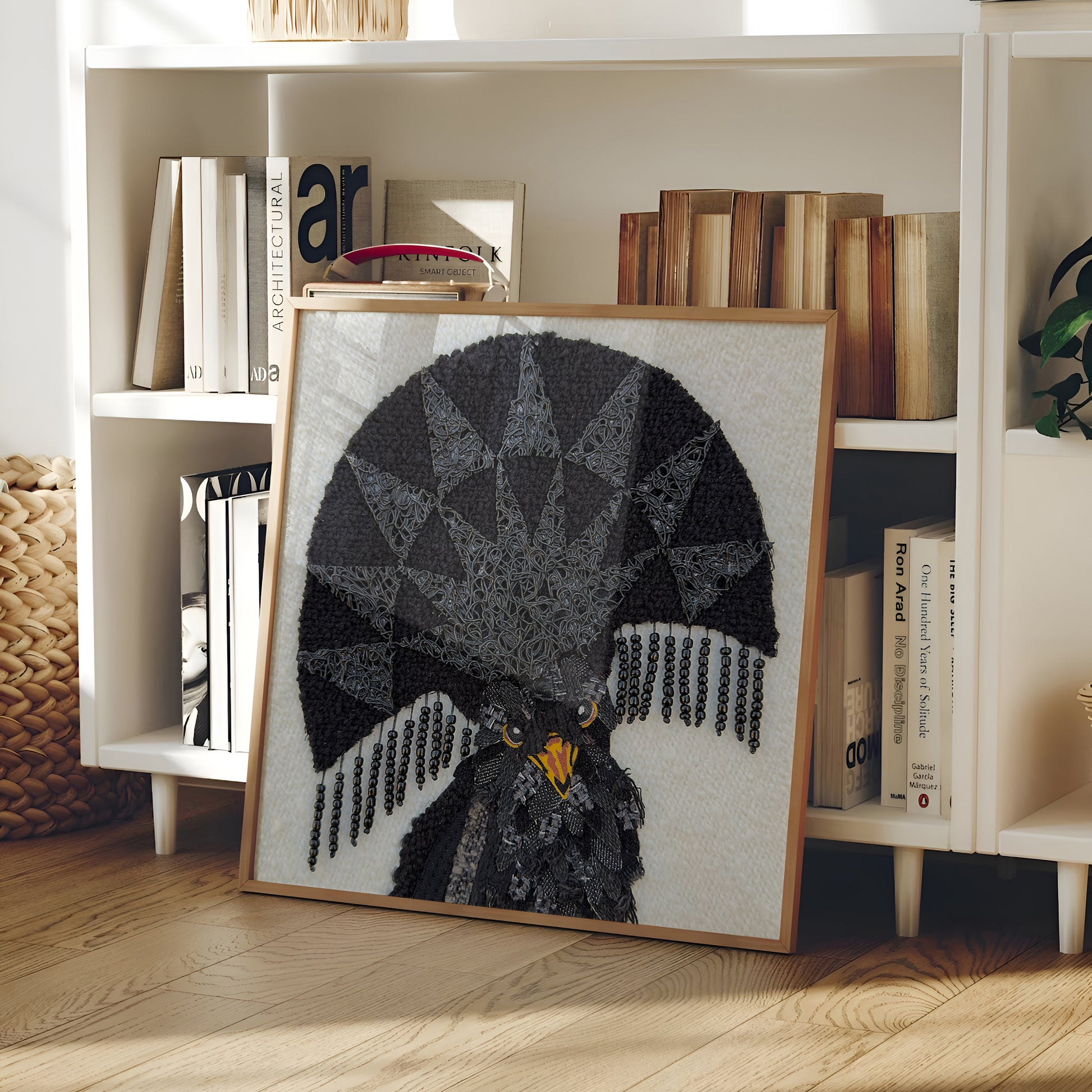 A framed needlepoint artwork of a black eagle with a geometric fan-shaped design and yellow eyes, leaning against a white bookshelf in a home interior.