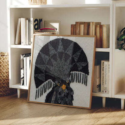 A framed needlepoint artwork of a black eagle with a geometric fan-shaped design and yellow eyes, leaning against a white bookshelf in a home interior.