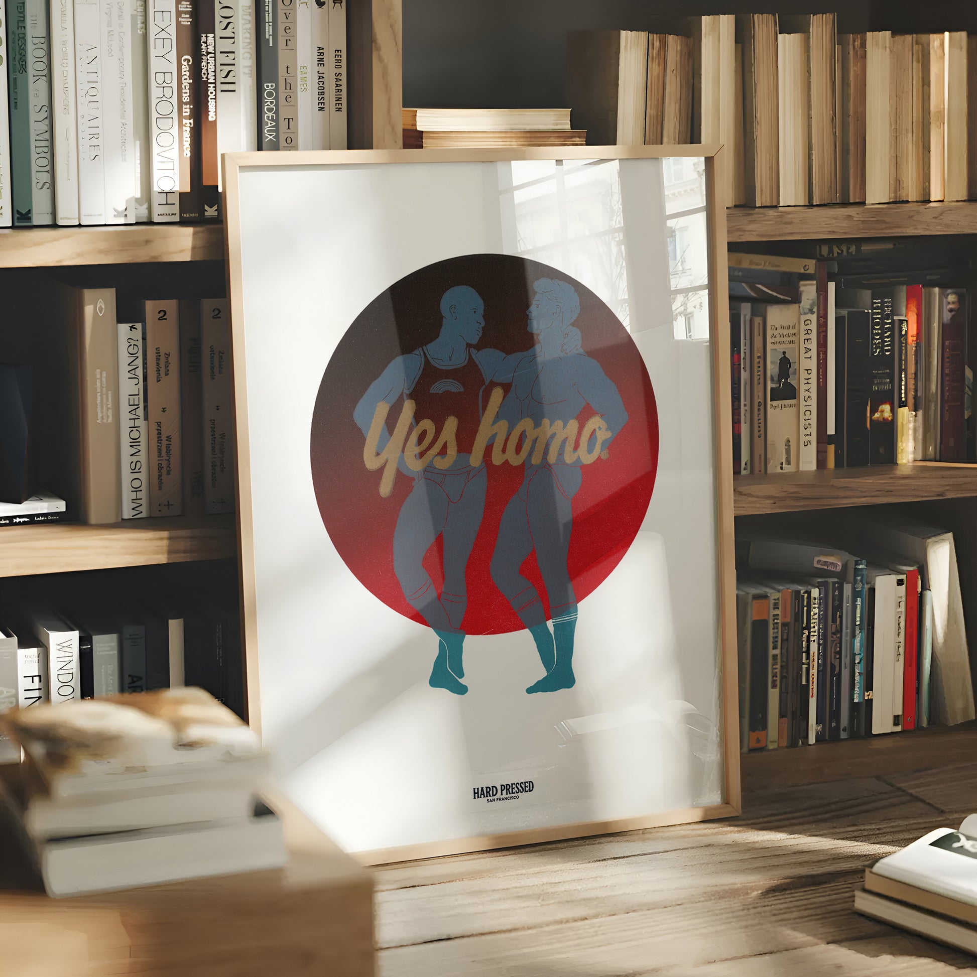 A framed art print leaning on a wooden table in front of a bookshelf. The print features a minimalist graphic of two dancing figures in teal and blue inside a large red circle with the text 'Yes homo' in gold.