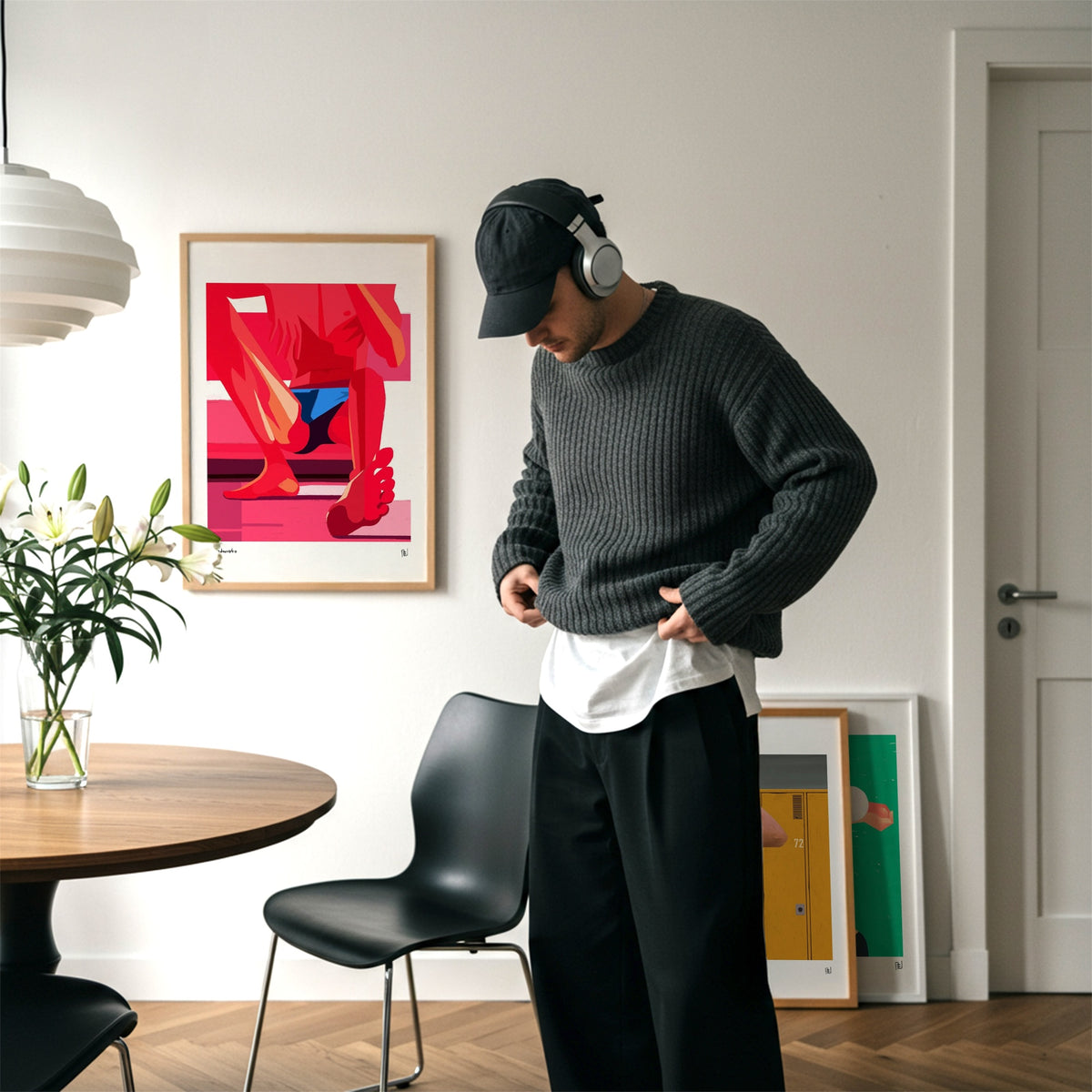 A man wearing a dark gray ribbed knit sweater, black pants, a black cap, and white headphones stands in a modern room, holding up the hem of his sweater to show a white undershirt. A framed abstract art print hangs on the wall behind him.