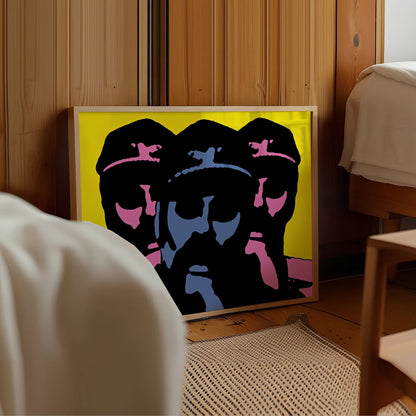 A framed pop-art print leaning against a wooden wall in a bedroom setting. The artwork features three stylized faces in black, blue, and pink on a bright yellow background, with a light wood frame.