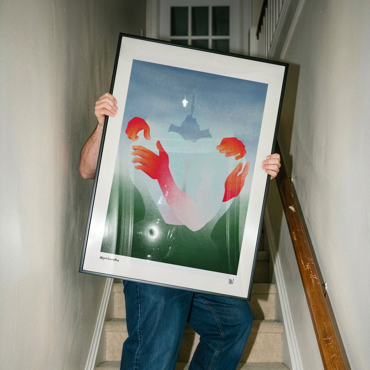 A person standing on an indoor staircase holds up a large framed art print. The artwork shows a surreal scene with red and pink hands reaching toward a glowing light in a blue-green landscape.