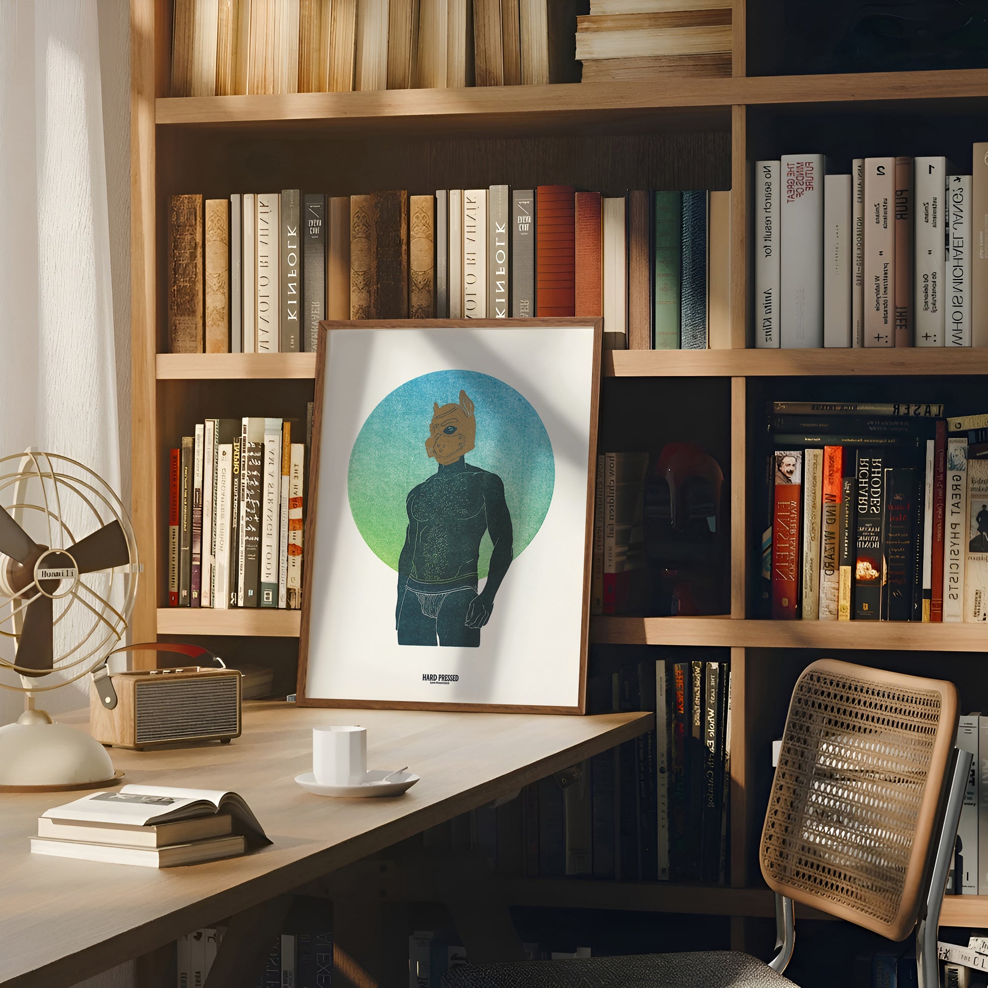 A framed art print featuring a stylized graphic portrait of a man with a pup mask on a teal and blue circular background, displayed on a wooden desk in front of a bookshelf.