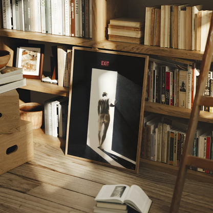 A framed black and white art print leaning on a wooden floor in a cozy, sunlit room filled with bookshelves and books. The print features a minimalist figure walking into a doorway with a red EXIT sign at the top.