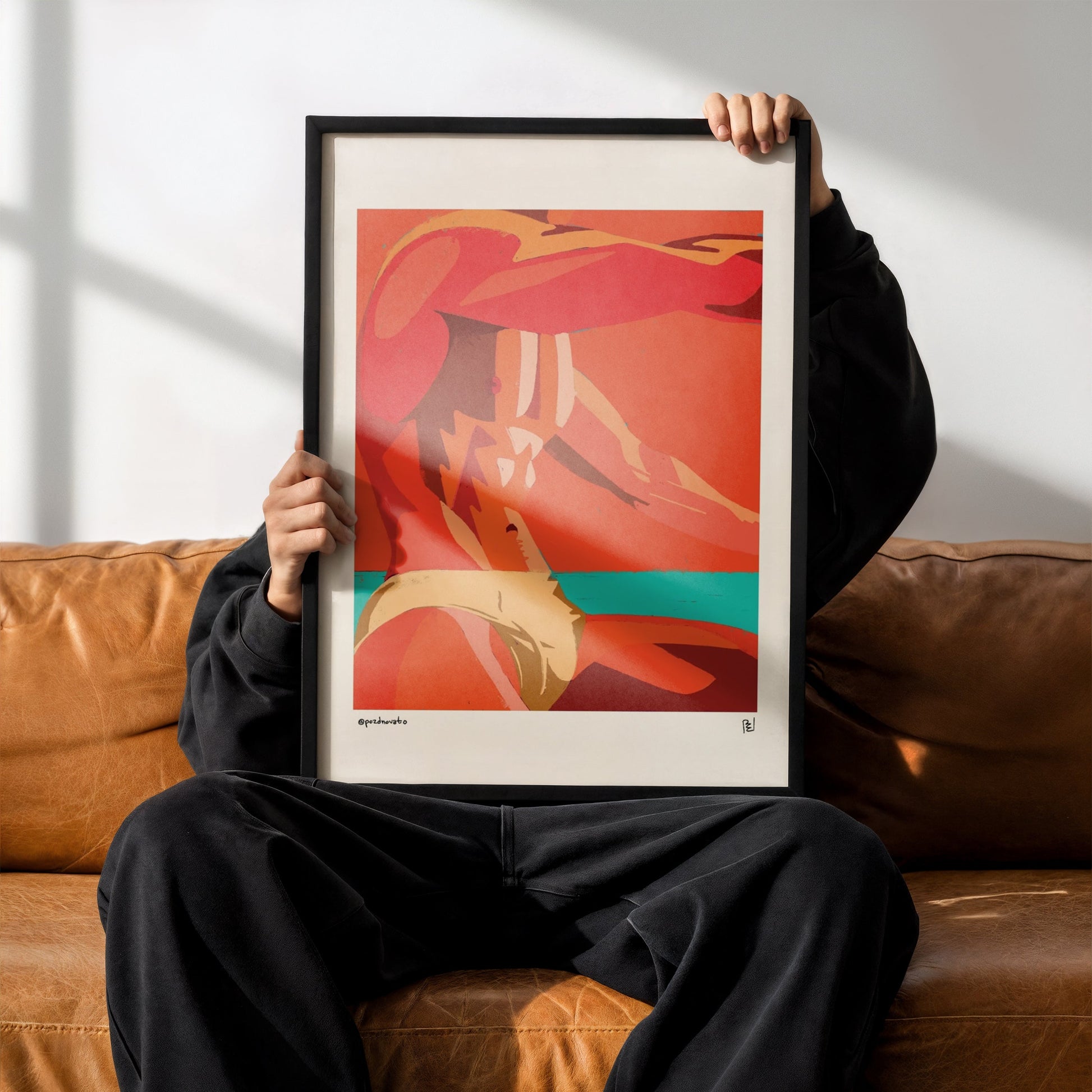 A person sitting on a brown leather couch holds up a framed abstract art print. The print features a bold composition of red, orange, and beige tones with a teal accent, set in a black frame.