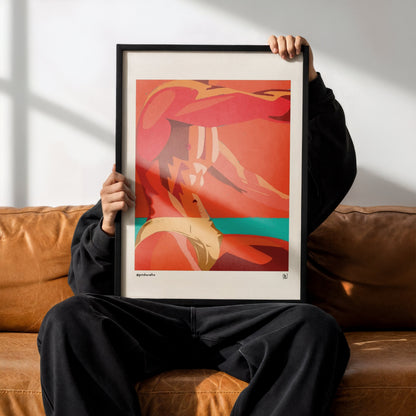 A person sitting on a brown leather couch holds up a framed abstract art print. The print features a bold composition of red, orange, and beige tones with a teal accent, set in a black frame.