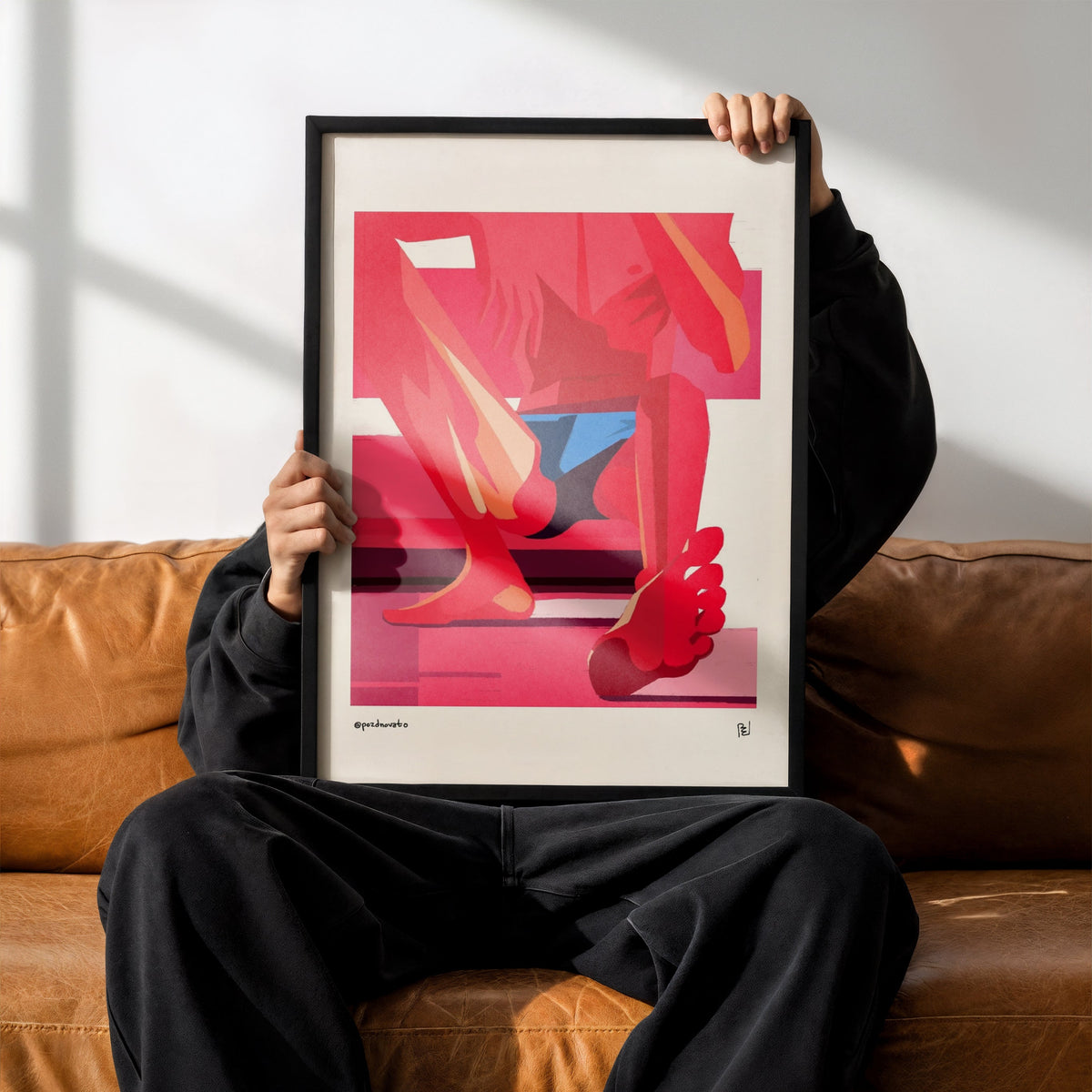 A person sitting on a brown leather couch holds up a framed art print featuring a stylized illustration of legs and feet in pink, red, and blue tones. The person is wearing a black hoodie, black sweatpants, and black Adidas sneakers.