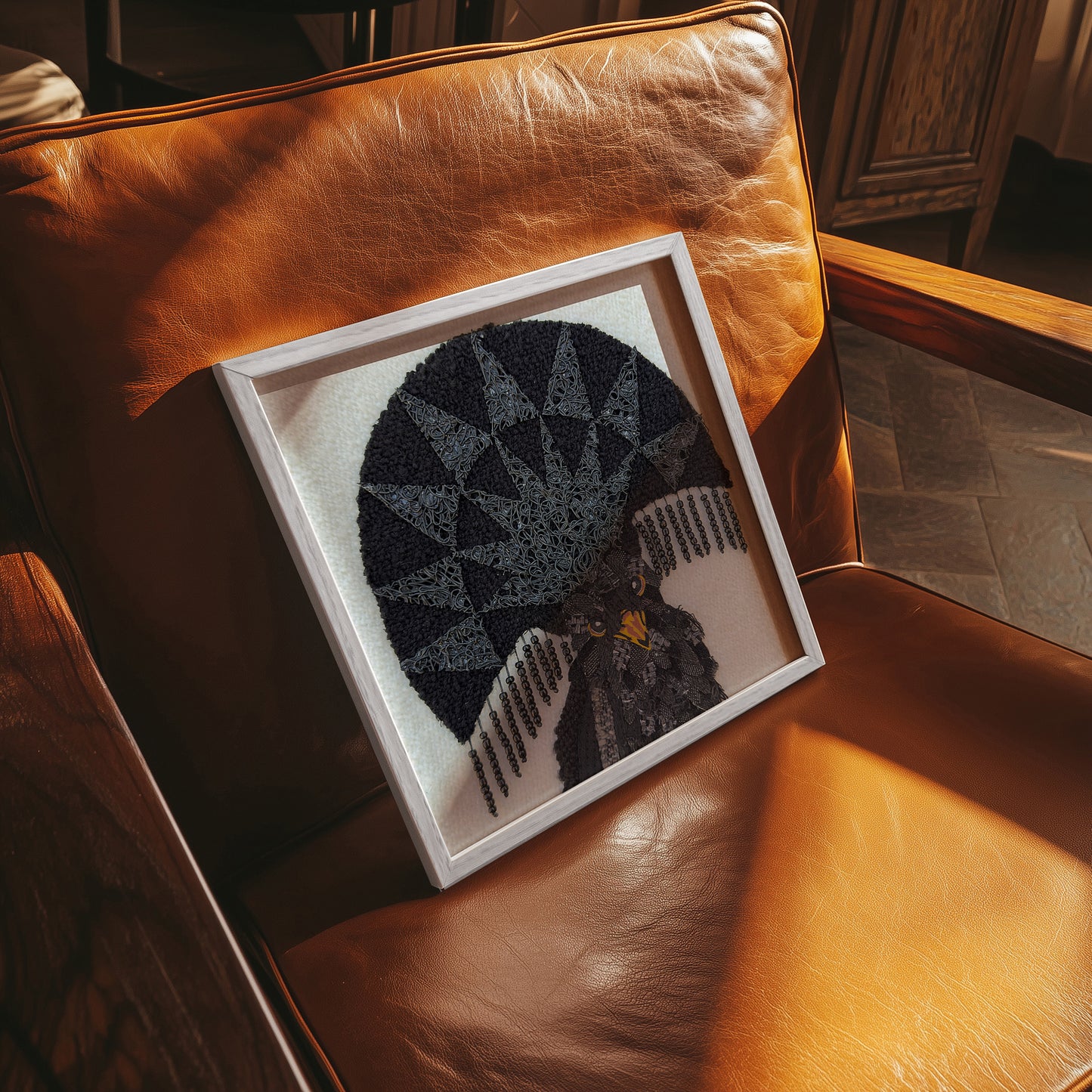 A framed needlepoint artwork featuring a stylized bird with a geometric fan motif, resting on a warm brown leather armchair in a sunlit room.