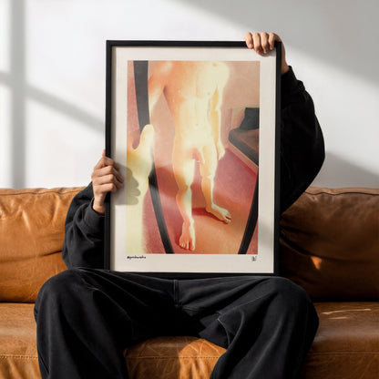 A person sitting on a brown leather couch holds up a framed art print featuring a stylized, warm-toned illustration of a nude human figure. The person is wearing a black hoodie, black sweatpants, and black Adidas sneakers with white stripes.
