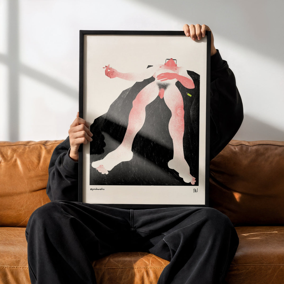 A person sitting on a brown leather couch holds up a framed art print featuring a stylized figure in pink, black, and white. The person is wearing a black hoodie, black sweatpants, and black Adidas sneakers with white stripes.