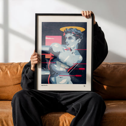 A person sitting on a brown leather couch holds up a framed art print featuring a stylized illustration of a woman with a halo and red geometric accents. The person is wearing a black hoodie, black sweatpants, and black Adidas sneakers.