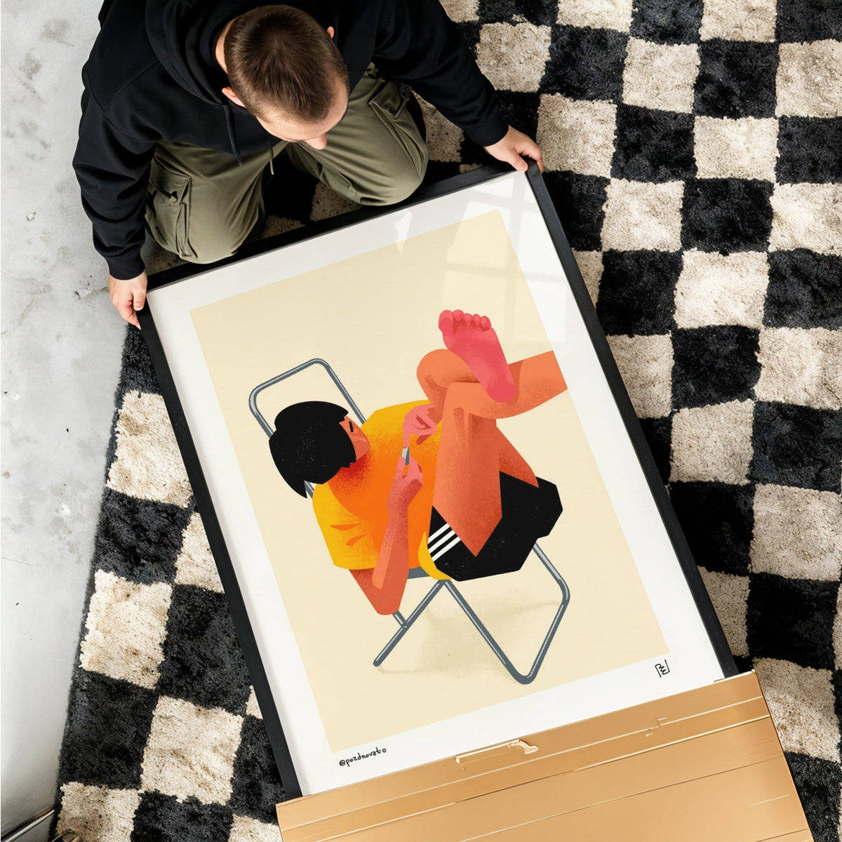 A framed art print featuring a stylized illustration of a person sitting on a chair, held by a person on a black and white checkered rug. The print is shown in its cardboard shipping box, with a gray concrete floor surrounding the scene.
