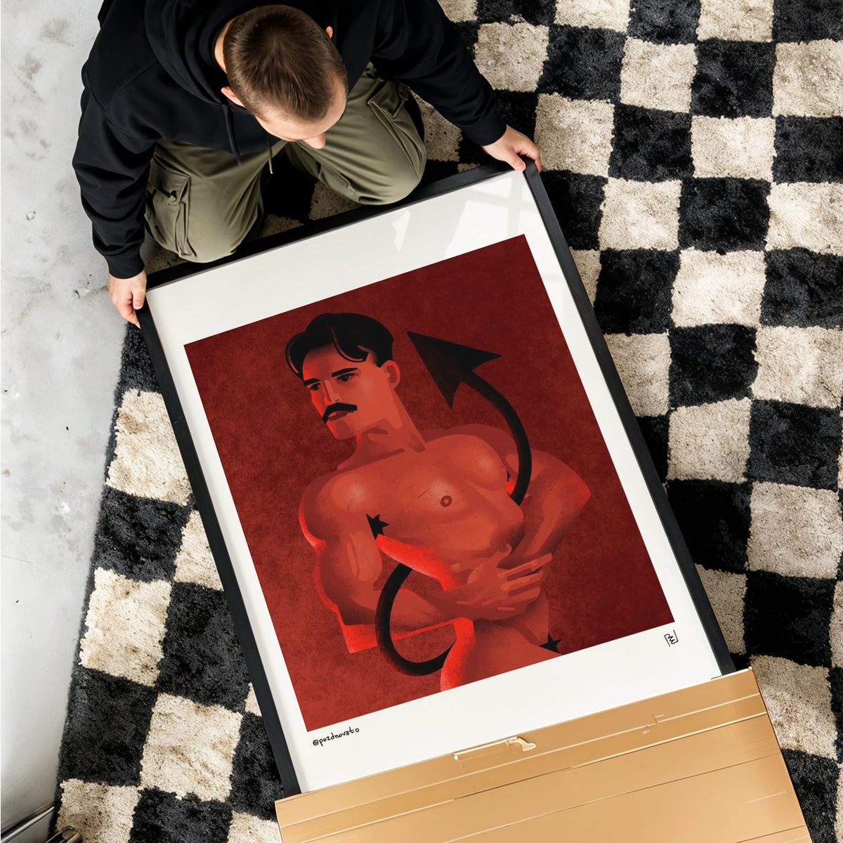 A framed art print featuring a muscular shirtless man with a mustache against a red background, held by a person on a black and white checkered rug. A large cardboard box is visible in the foreground.