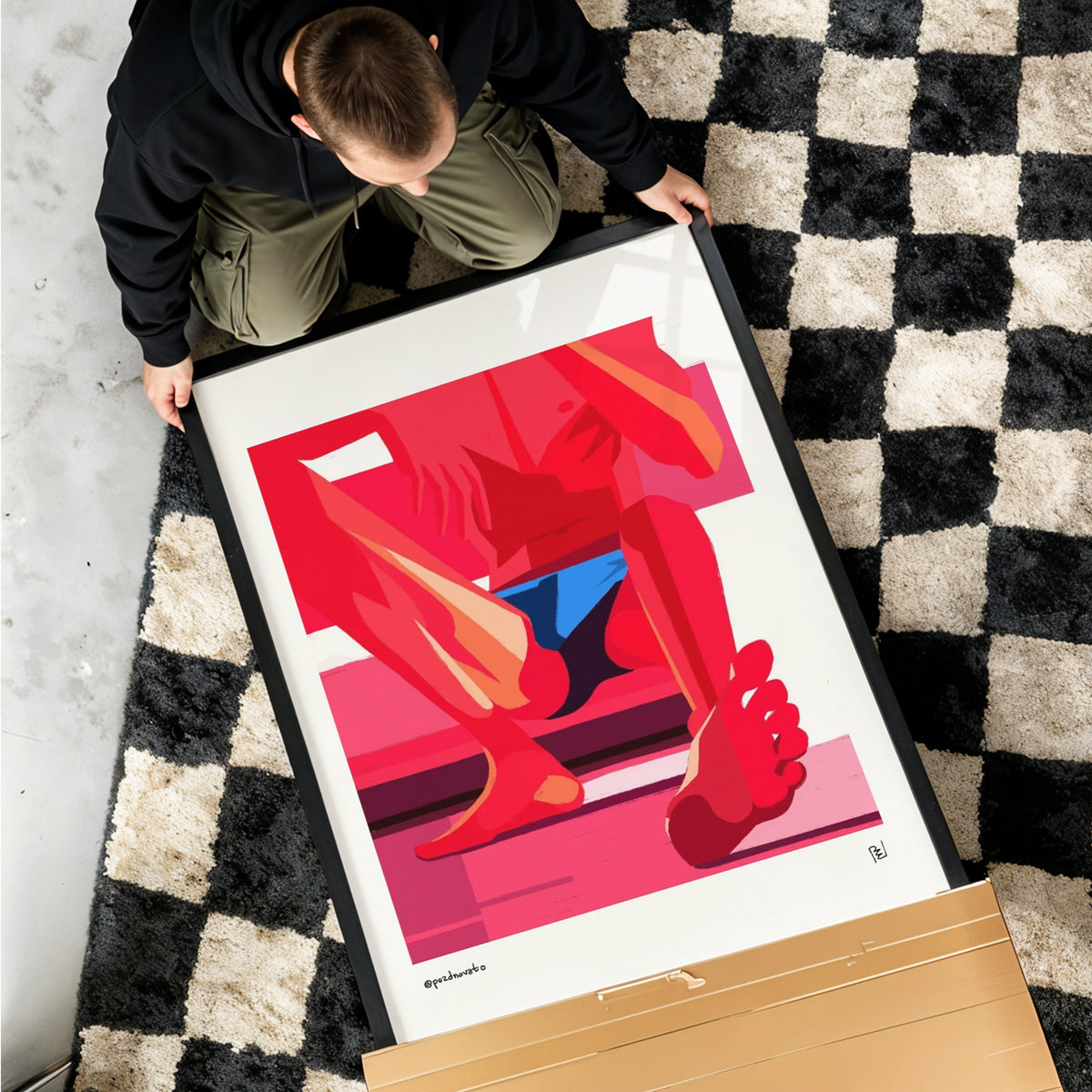 A framed art print featuring a vibrant red and pink abstract illustration of legs and feet, held by a person on a black and white checkered rug. A large cardboard box lies on the floor nearby.