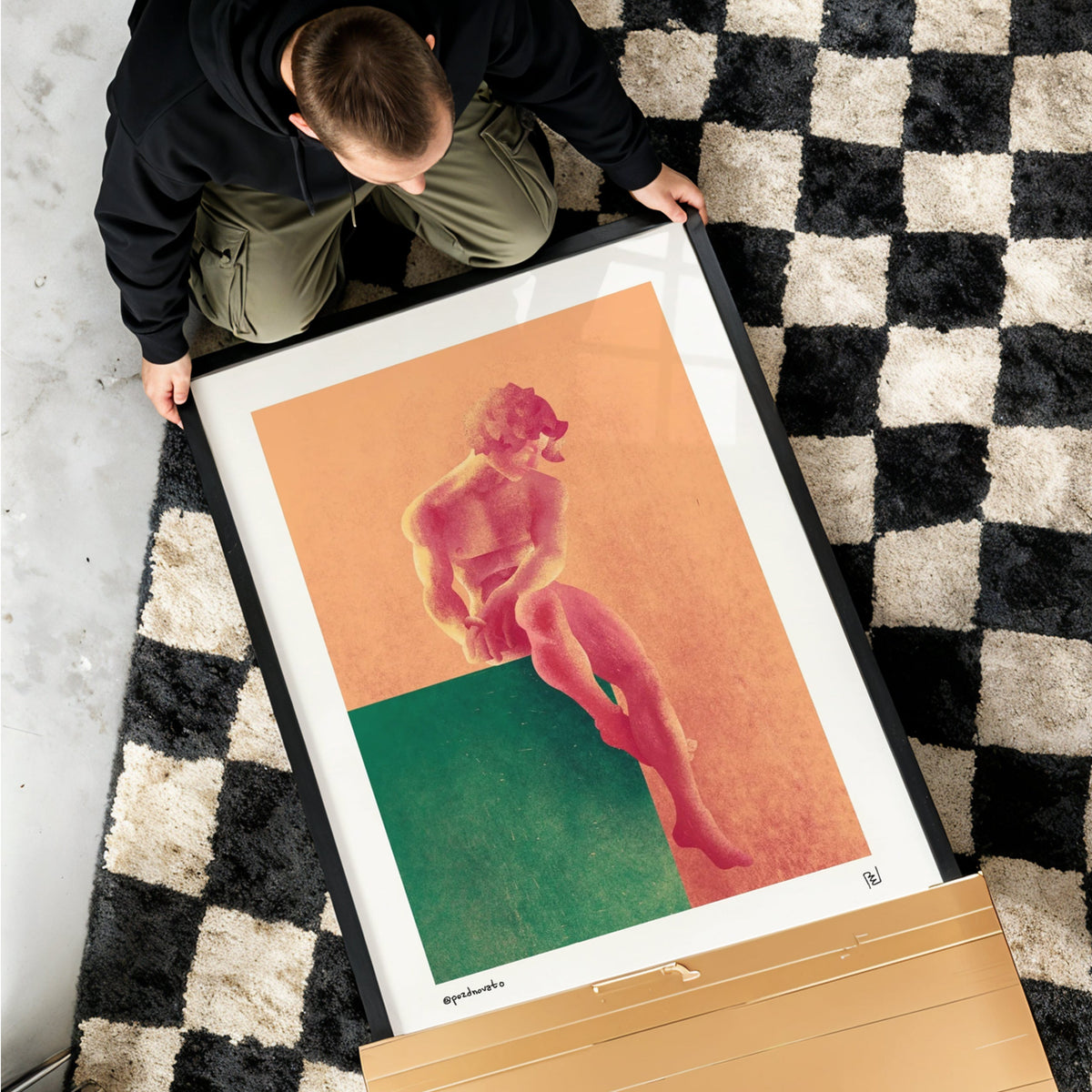 A framed art print featuring a stylized pink figure against peach and green blocks, held by a person on a black-and-white checkered rug. The print is shown in a real indoor setting with a cardboard box nearby.