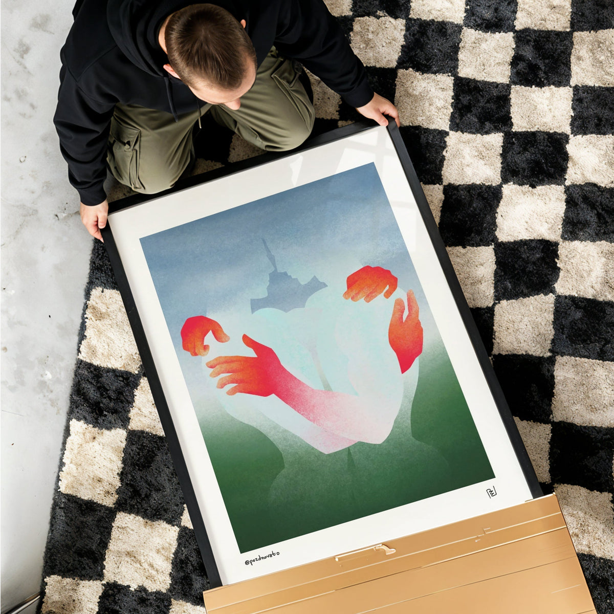 A person kneels on a black and cream checkered rug while handling a large framed art print. The print features a minimalist illustration of two hands reaching toward a small tower, with a light blue sky and green ground.
