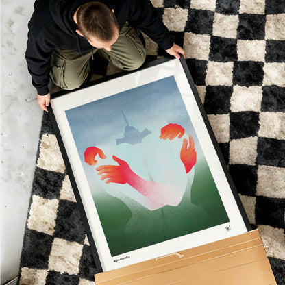 A person kneels on a black and cream checkered rug while handling a large framed art print. The print features a minimalist illustration of two hands reaching toward a small tower, with a light blue sky and green ground.