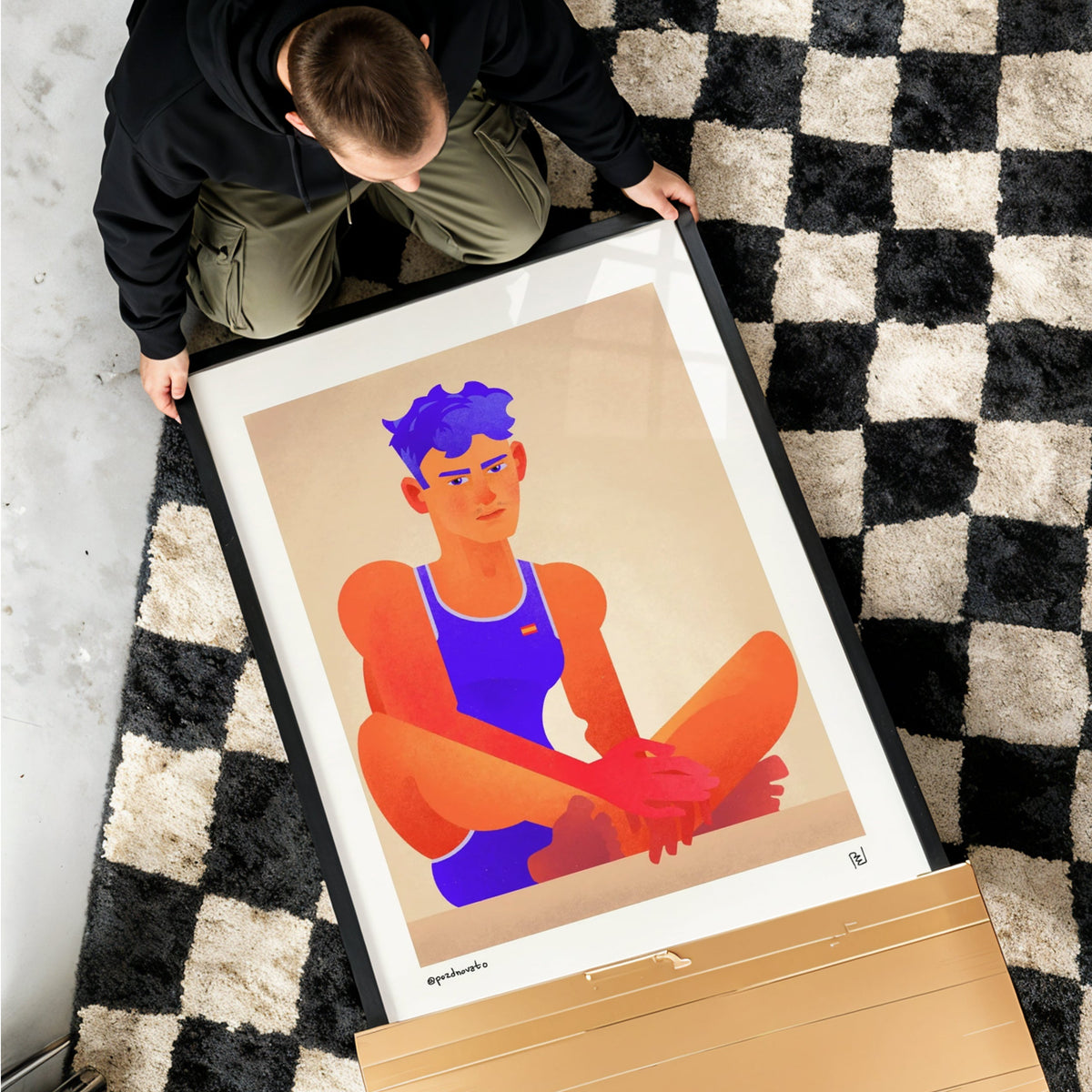 A large framed art print featuring a stylized illustration of a person in a blue tank top and orange pants, held by a person on a black and white checkered rug. A cardboard box is visible in the foreground.