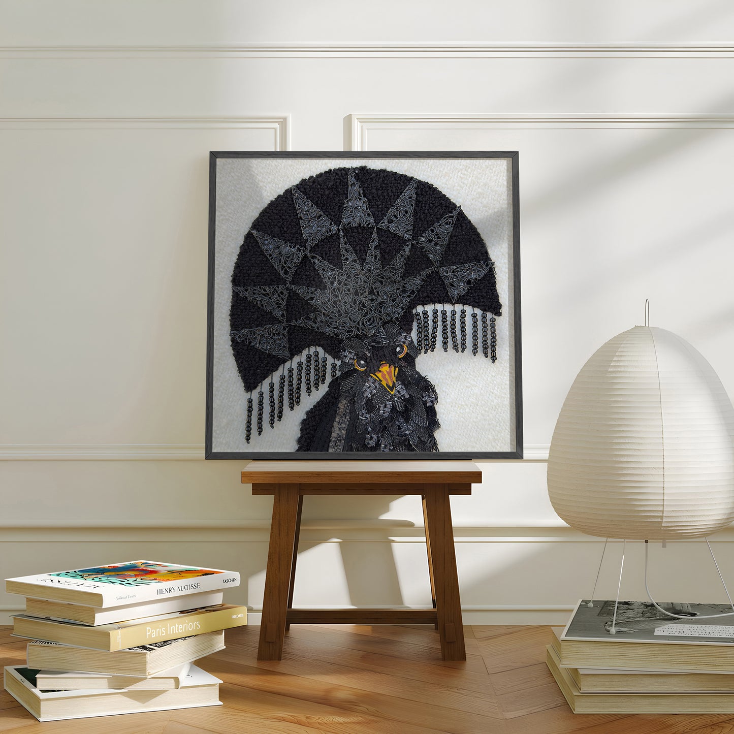 A framed art print of a black bird with a large fan-like crest, displayed on a wooden stool in a styled room with books and a lamp.