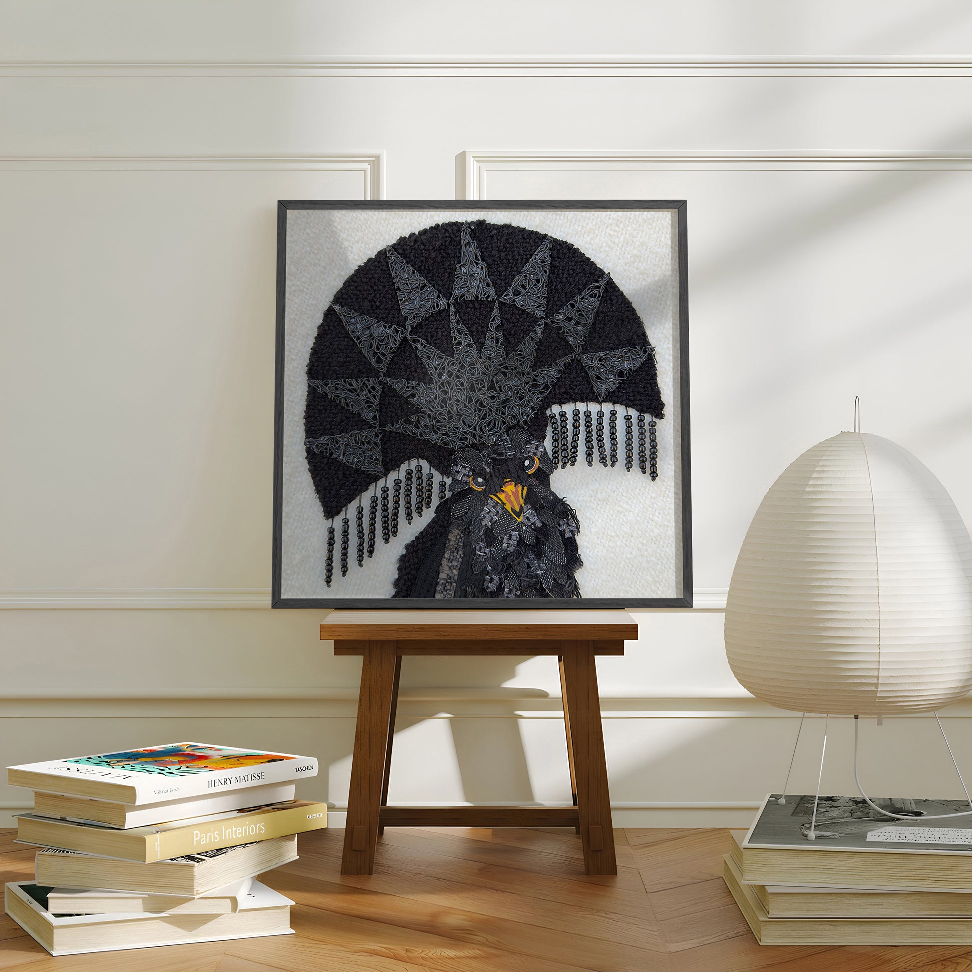 A framed art print of a black bird with a large fan-like crest, displayed on a wooden stool in a styled room with books and a lamp.