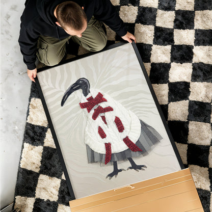 A person kneels on a black and cream checkered rug while holding a large framed art print featuring a stylized bird in a white coat with red accents. A light wood panel lies in the foreground on a gray floor.