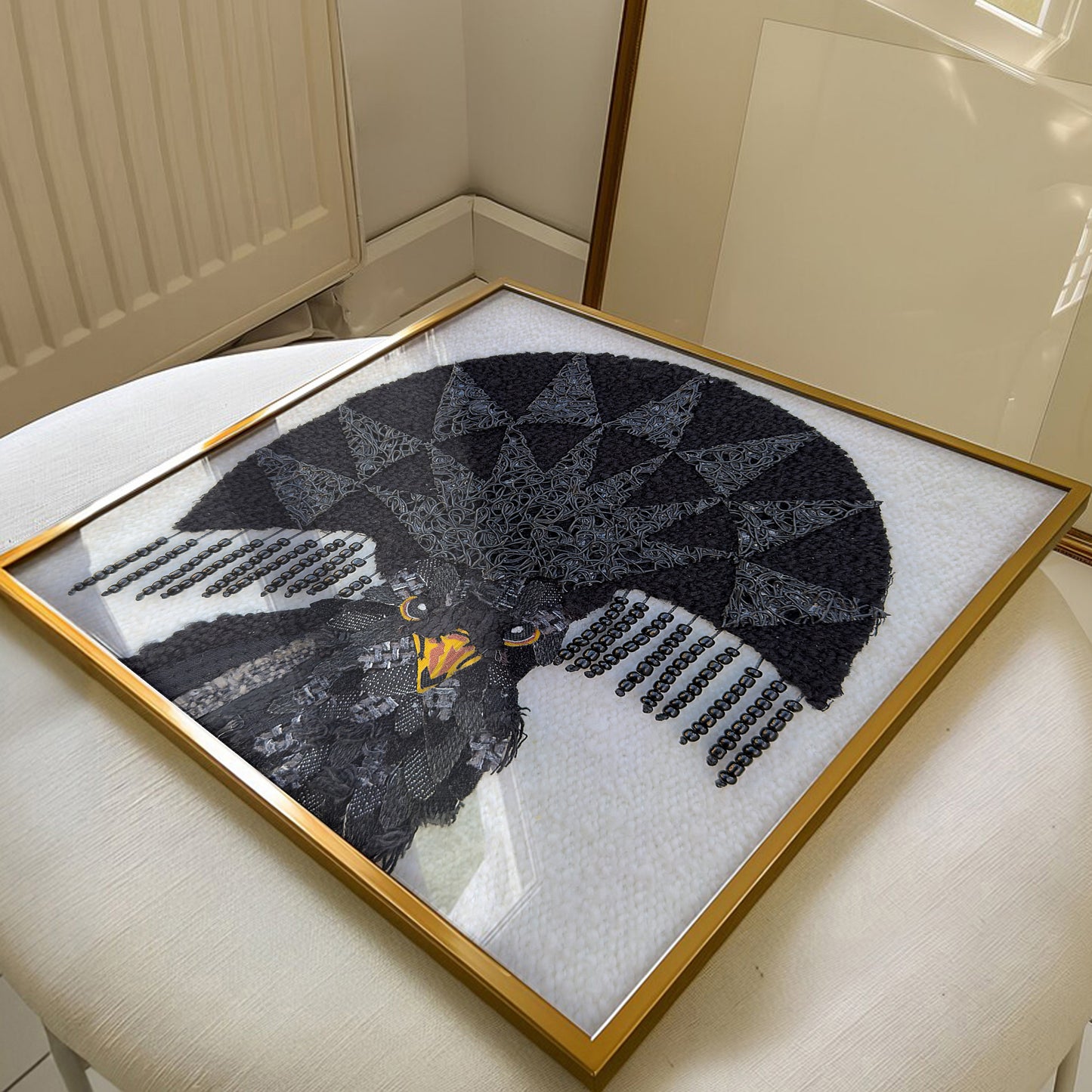 A framed black lace needlework artwork of a bird with geometric wing patterns, displayed on a light-colored upholstered chair in an indoor setting.