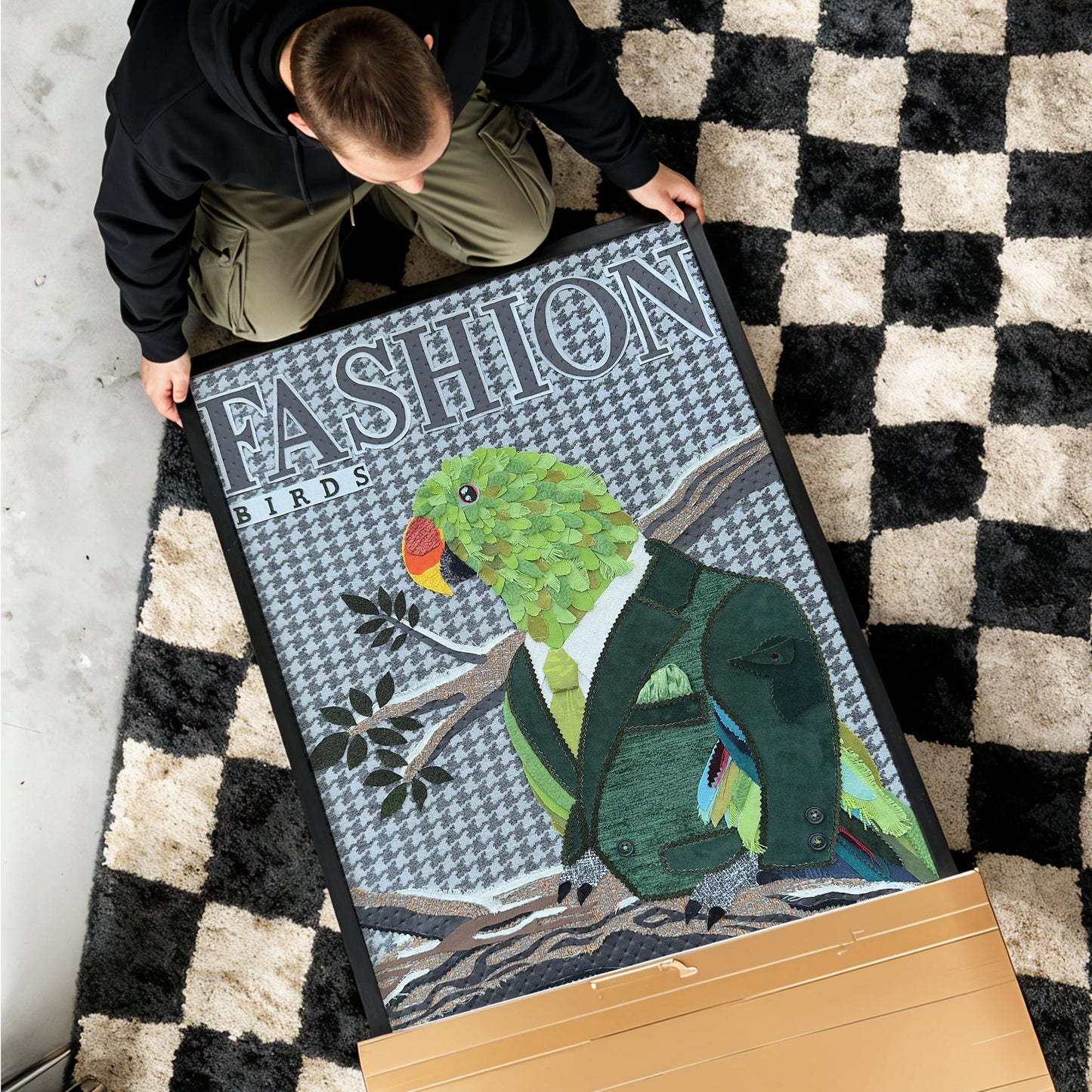 A person is kneeling on a black and cream checkered rug, holding a large framed art print featuring a green parrot in a dark green suit with the word 'FASHION' on the frame. A large cardboard box is placed in the foreground on a light gray concrete floor.