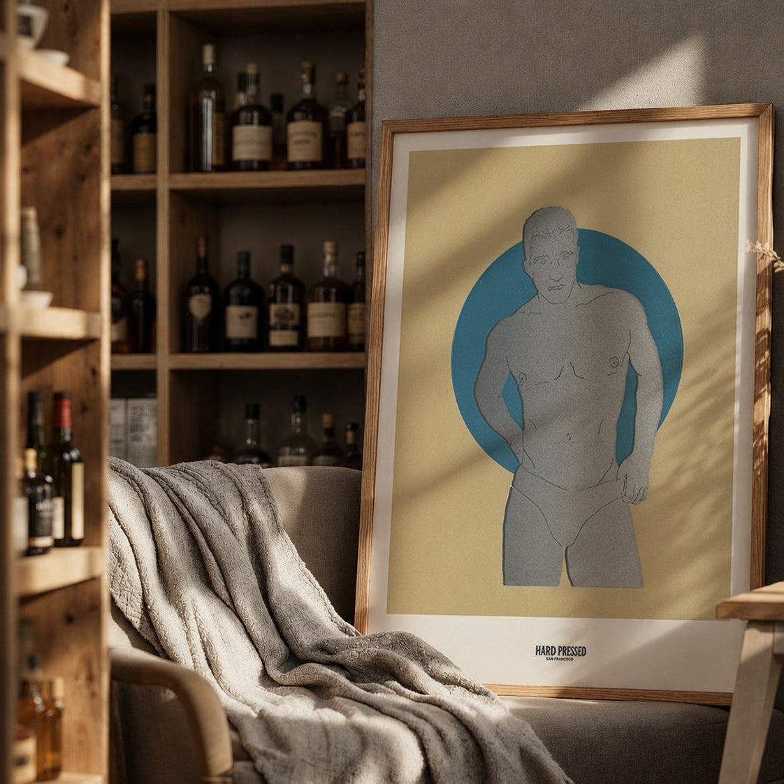 A framed art print featuring a minimalist gray male figure silhouette against a blue circular background on a cream-colored print, styled in a warm, rustic room next to a wooden shelf of bottles and a cozy armchair.