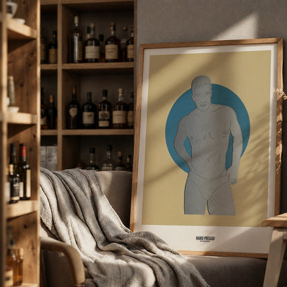 A framed art print featuring a minimalist gray male figure silhouette against a blue circular background on a cream-colored print, styled in a warm, rustic room next to a wooden shelf of bottles and a cozy armchair.