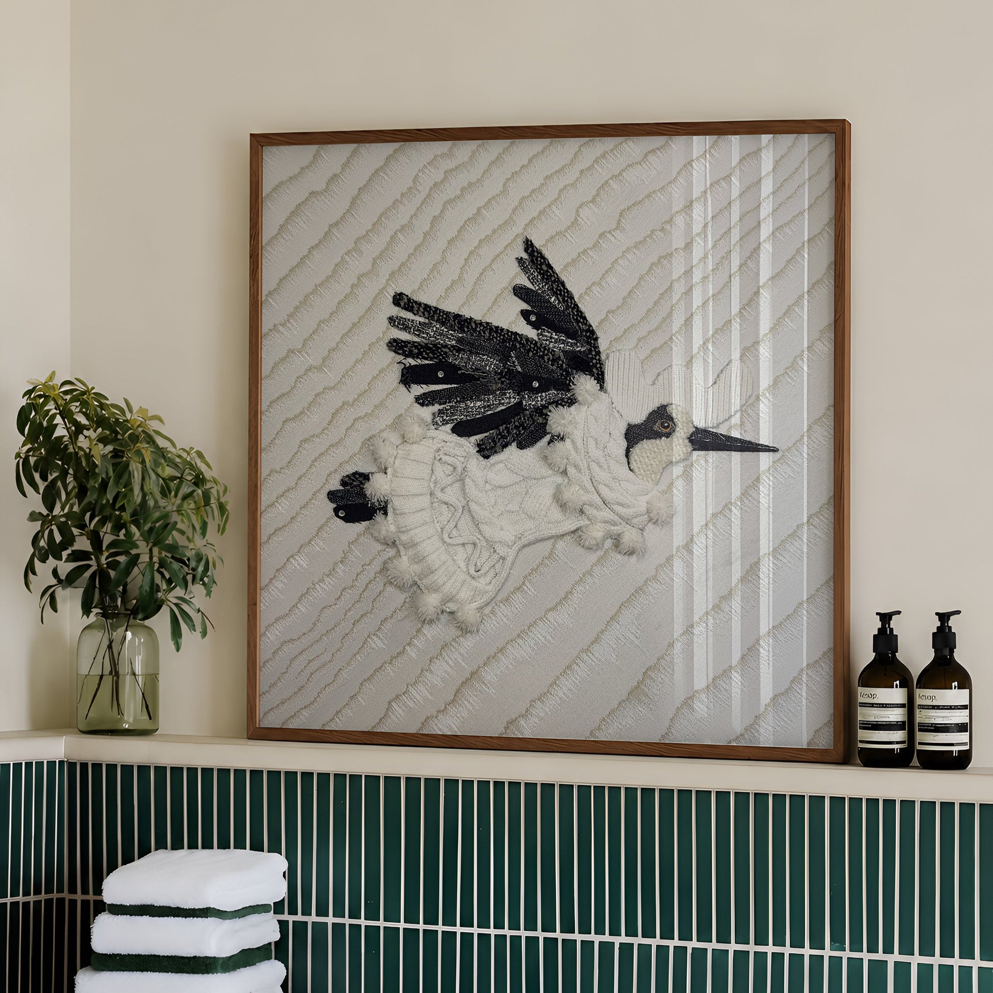 A framed wall art piece featuring a black-and-white bird sculpture on a textured cream background, displayed above a bathroom counter with green tile, folded towels, and two pump bottles.