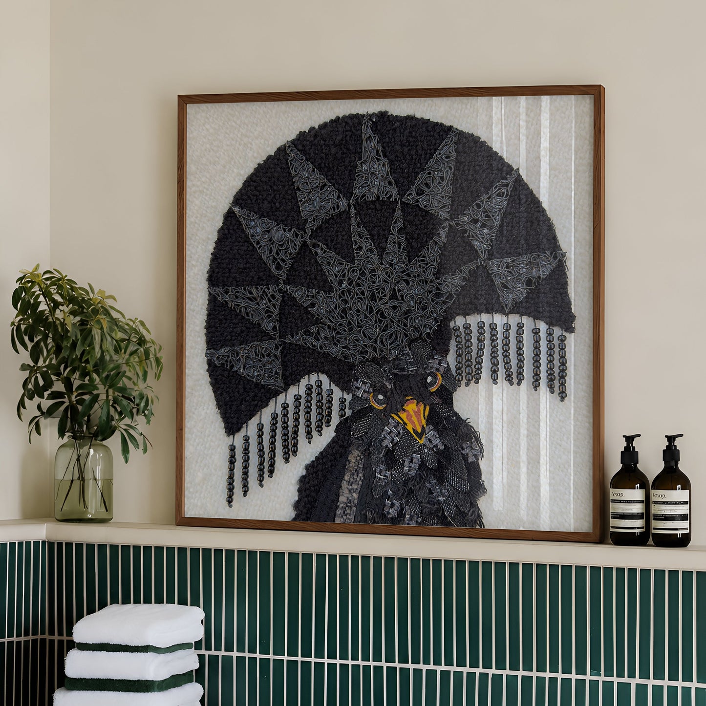 A framed art piece featuring a black owl with a large black fan-like geometric crest and yellow eyes, displayed on a bathroom shelf above a green-tiled counter with towels and toiletries.