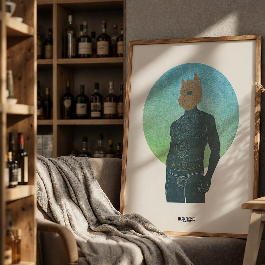 A framed art print featuring a stylized figure in dark blue against a blue and green circular background, leaning against a wall in a warm, rustic room with shelves of bottles, a chair, and dried flowers.