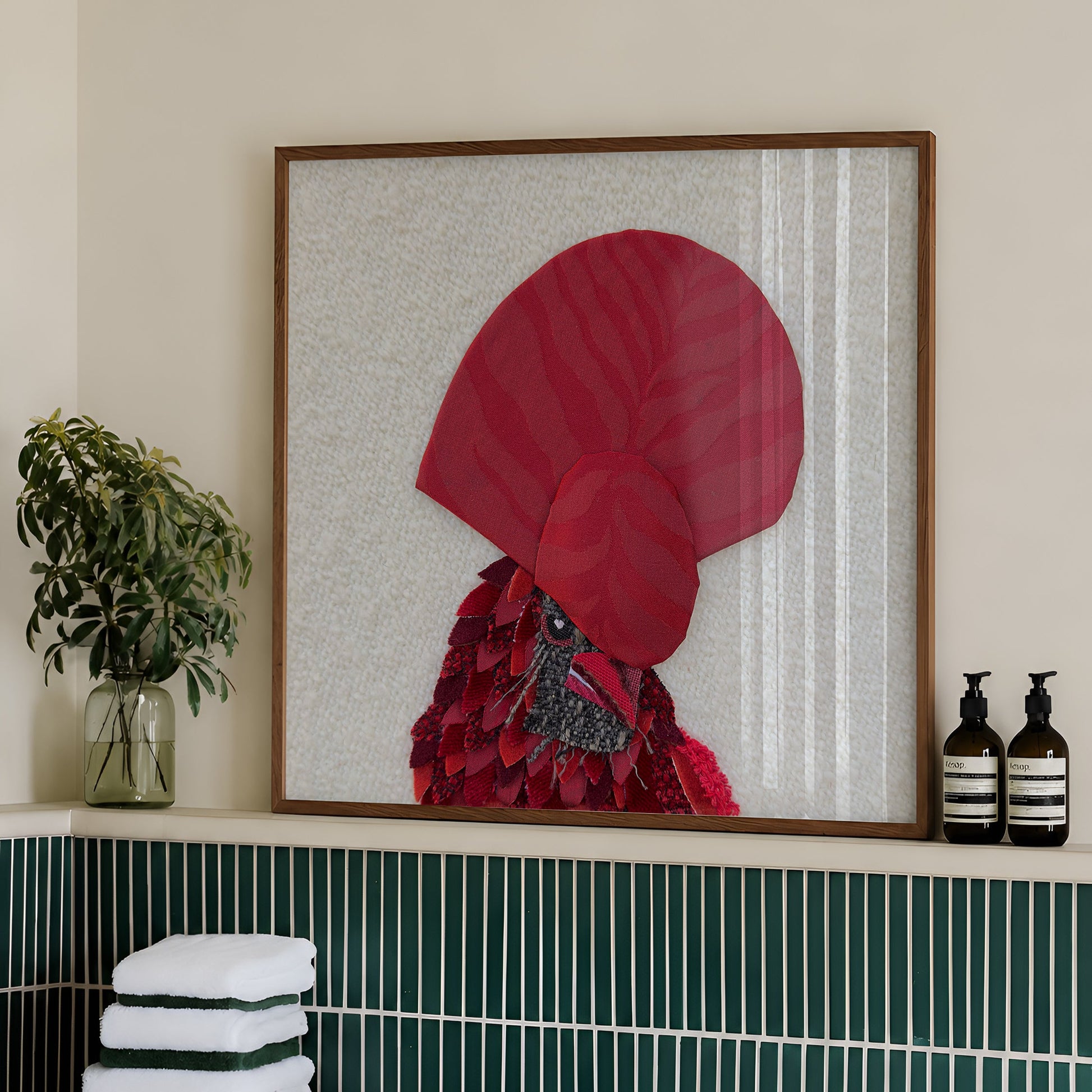 Framed wall art featuring a stylized red rooster with a large red comb, displayed in a bathroom setting above a green tiled ledge with folded white towels and toiletry bottles.