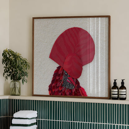 Framed wall art featuring a stylized red rooster with a large red comb, displayed in a bathroom setting above a green tiled ledge with folded white towels and toiletry bottles.