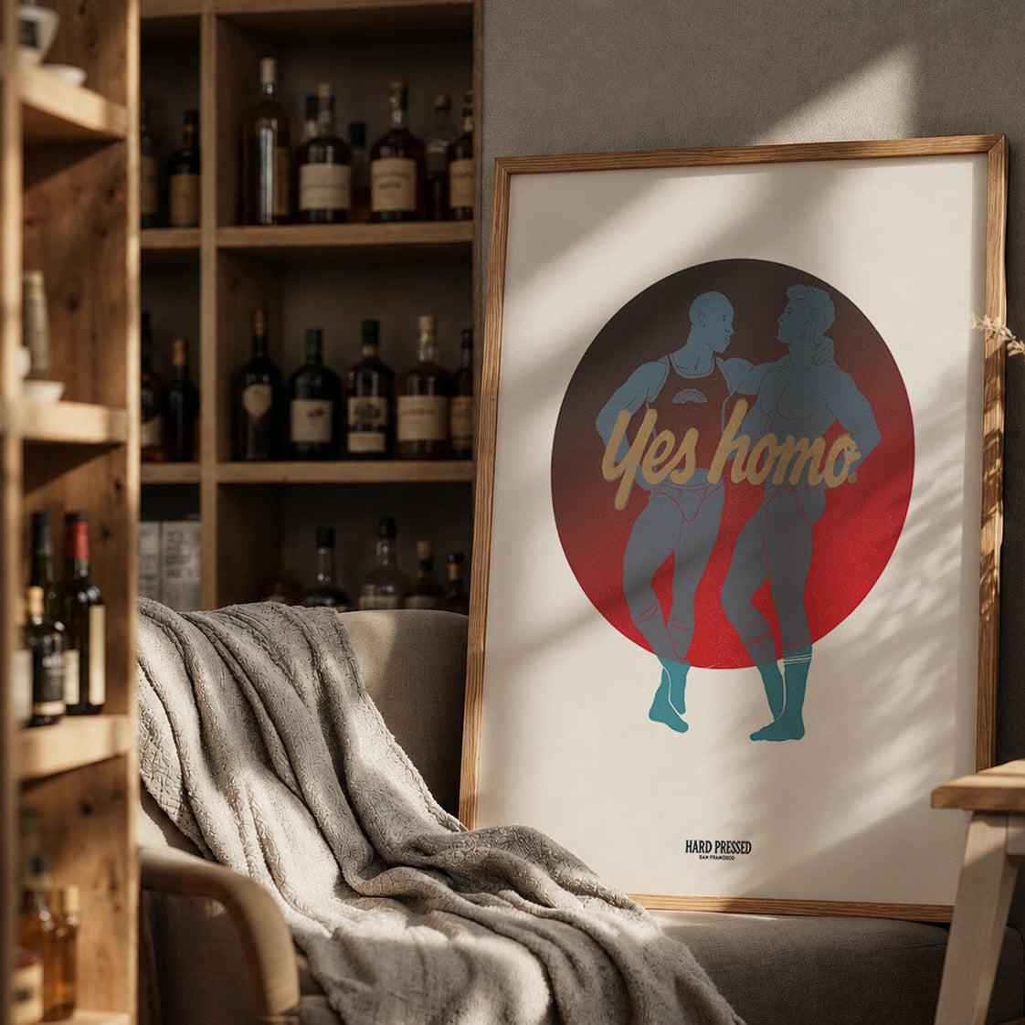 A framed art print leaning against a wall in a cozy room setting. The print features a minimalist design with the text 'Yes homo' in gold script over a red circle, with blue silhouettes of two people inside.
