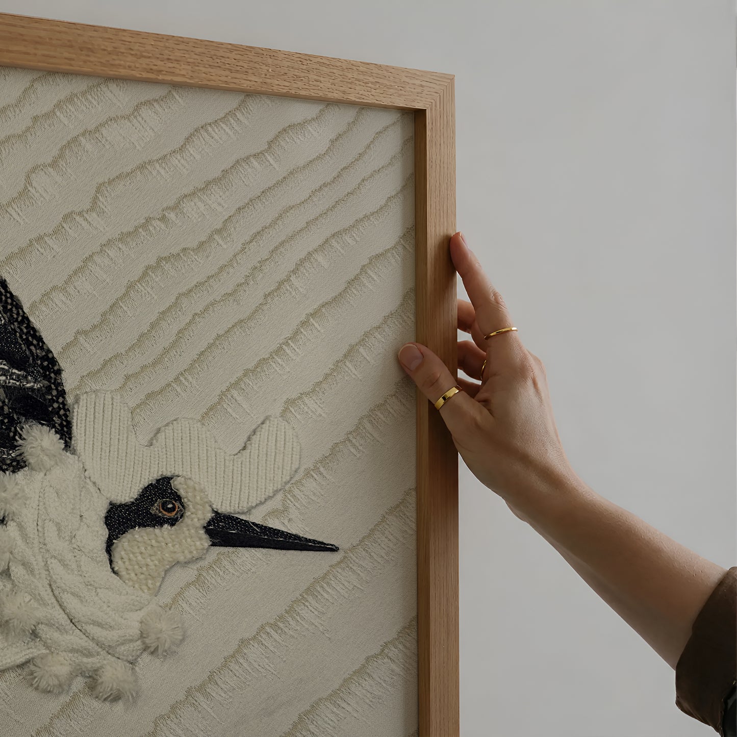 A close-up of a framed textile artwork being held by a person's hand. The artwork shows a black-and-white bird motif on a textured cream background, with a light natural wood frame visible against a plain light gray wall.