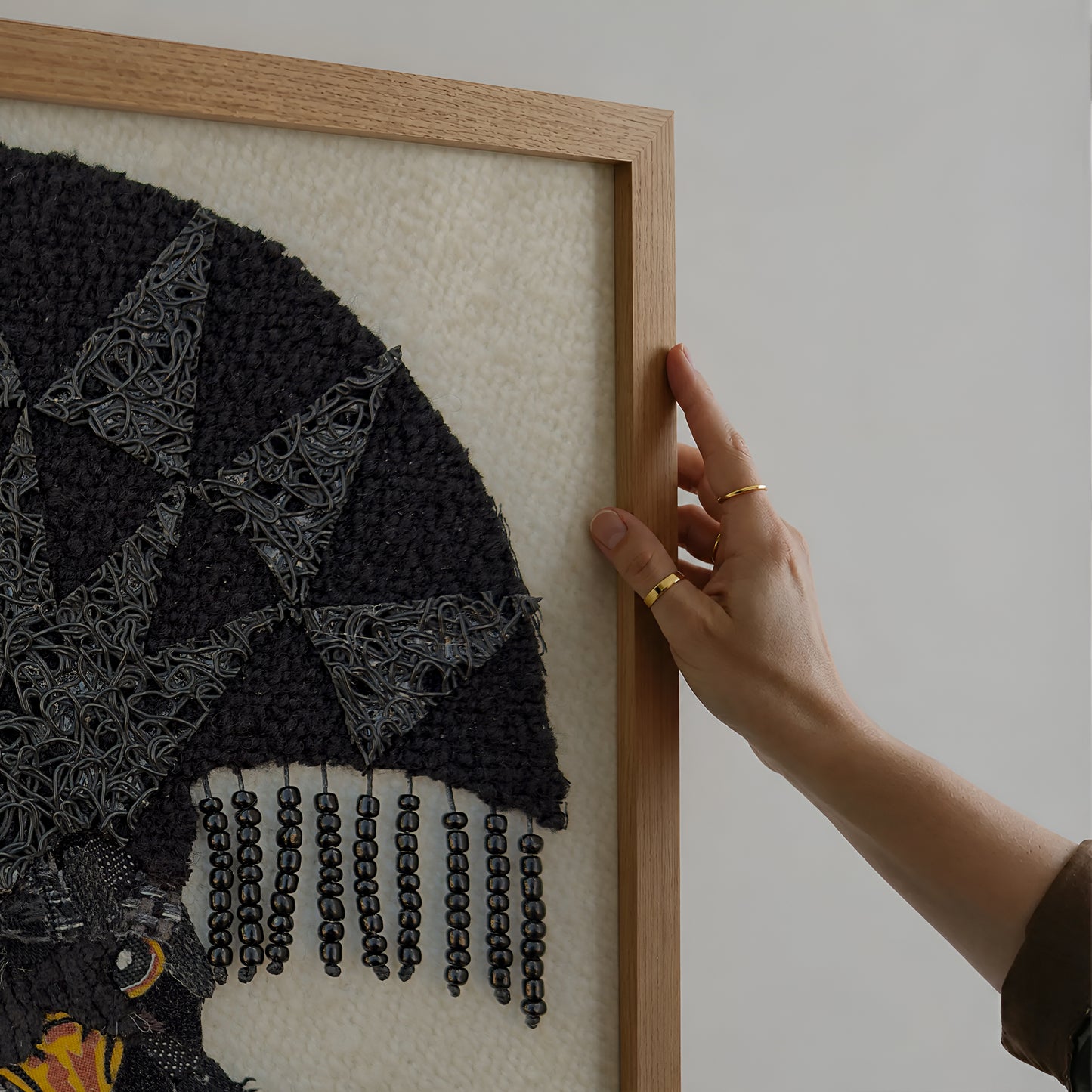 A close-up of a hand holding a light oak wooden frame next to a black and gray beaded wall art piece with a cream background.
