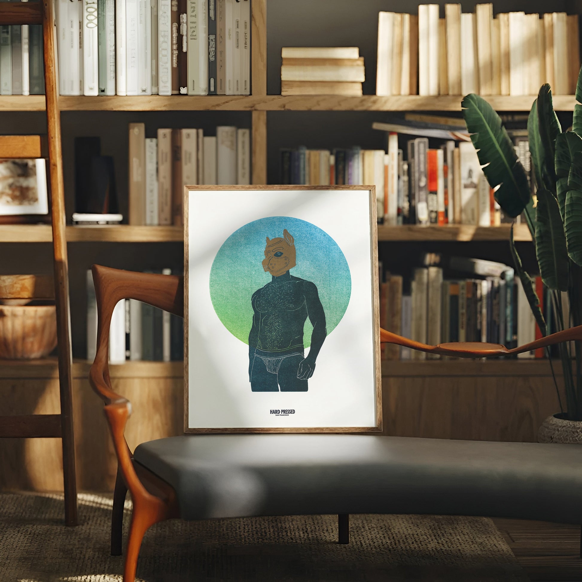 A framed art print featuring a stylized graphic portrait of a person with a pup mask inside a blue and green circular background, displayed on a gray bench in a cozy, sunlit book-filled room.