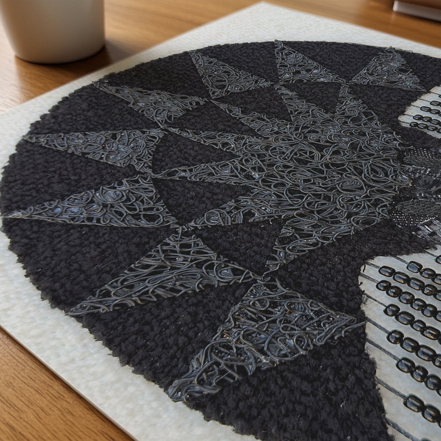 A close-up of a circular black and gray textured rug or mat with geometric star-like patterns, placed on a light surface over a wooden table. Part of a white mug is visible in the upper left.