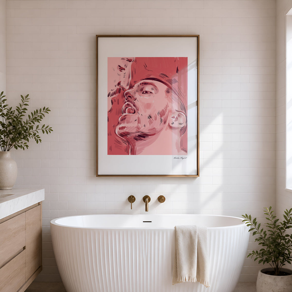 A framed art print featuring a pink-toned portrait hangs on a white brick wall above a freestanding white tub in a bright bathroom with plants and gold fixtures.