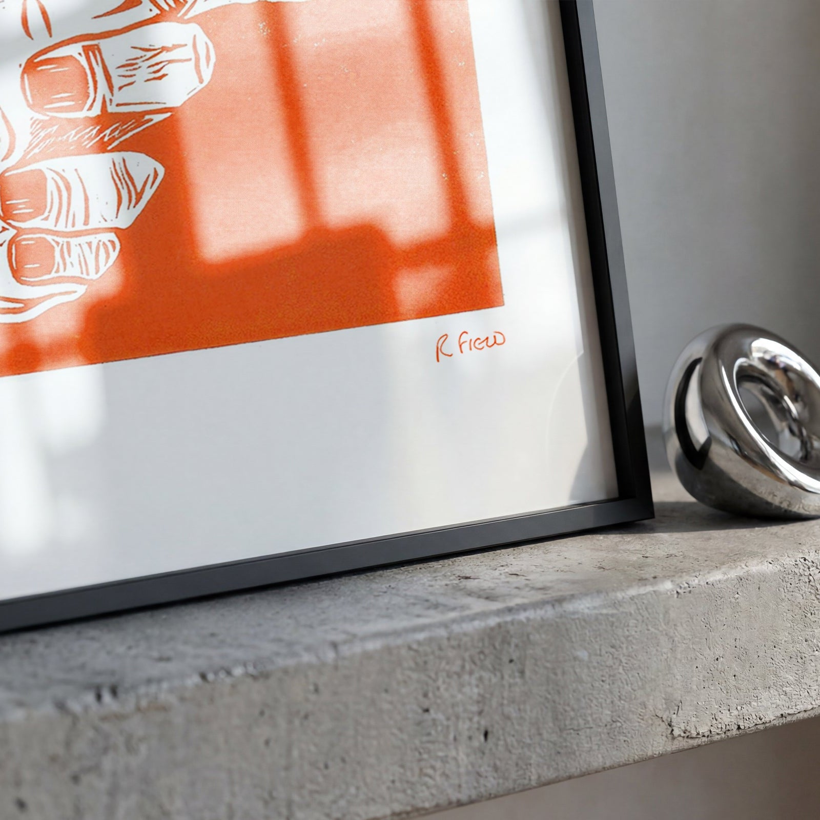 A framed art print featuring a stylized hand illustration in orange and white, leaning on a concrete ledge next to a polished silver ring.