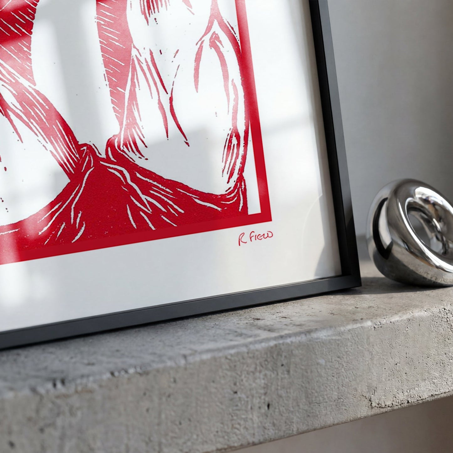 A framed art print with a bold red and white graphic design, resting on a textured concrete ledge next to a shiny silver metallic object.