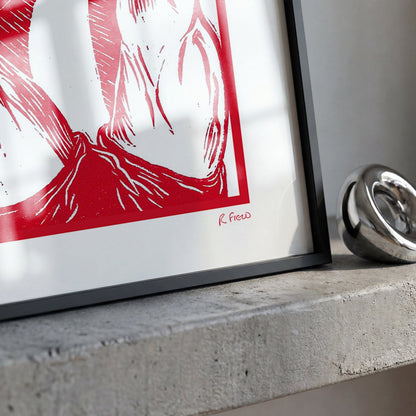 A framed art print with a bold red and white graphic design, resting on a textured concrete ledge next to a shiny silver metallic object.