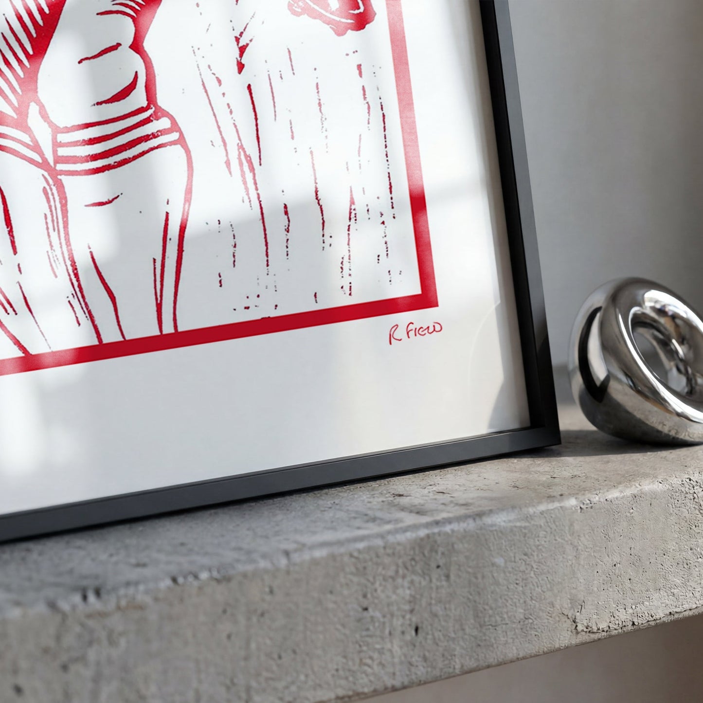 A framed art print with a bold red graphic design on a white background, leaning against a concrete ledge next to a polished silver metallic ring.