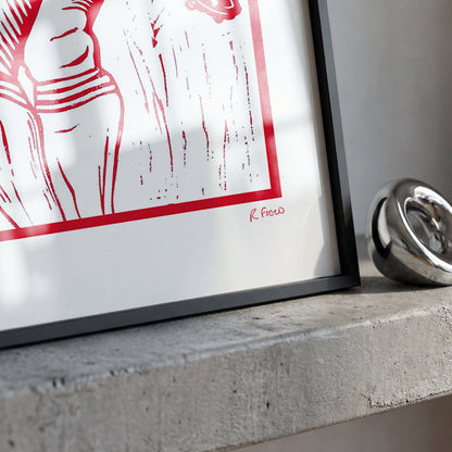 A framed art print with a bold red graphic design on a white background, leaning against a concrete ledge next to a polished silver metallic ring.
